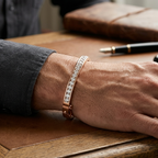 Hand wearing a bracelet on a wooden surface with a notebook and glass of whiskey in the background